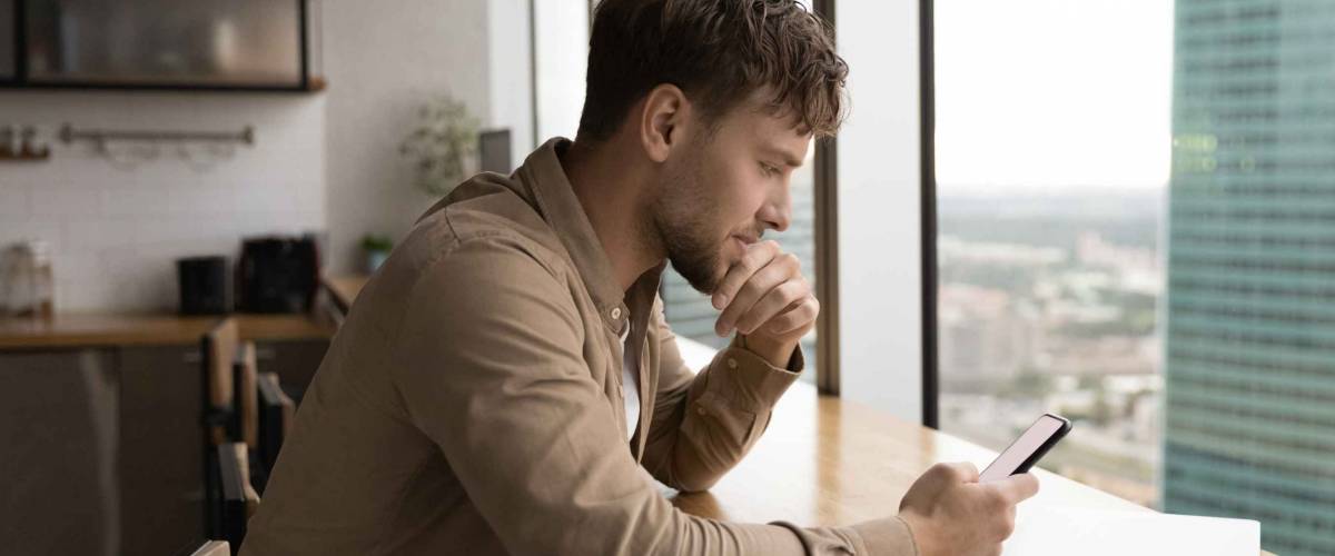 Information to think about. Pensive male look at cell ponder on job offer received by email. Young businessman sit by large window at modern kitchen, read important message on phone screen