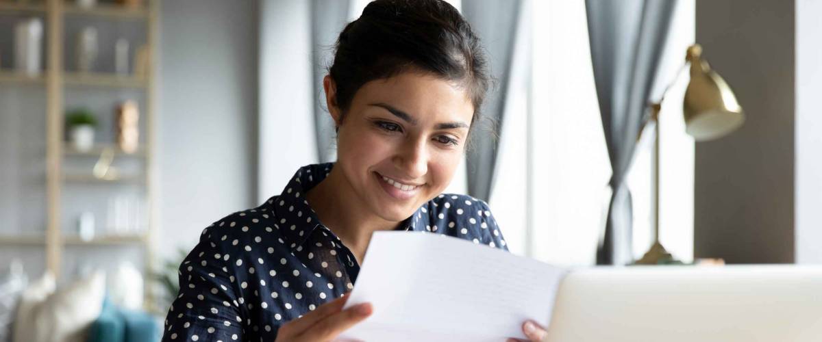 Smiling young woman holding letter, sitting in front of laptop in home office.