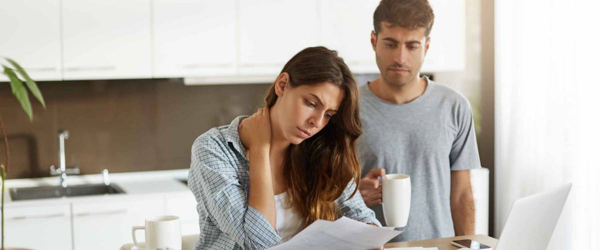 Stressed out couple sitting in kitchen, looking at bills and laptop.