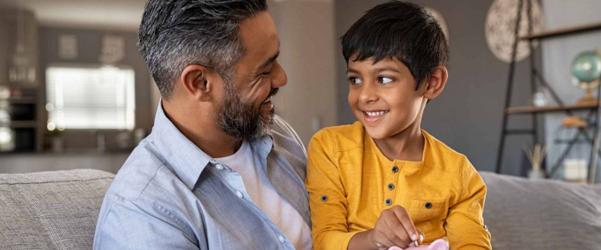 Little boy sitting on father's lap, smiling and putting money into a piggy. bank.