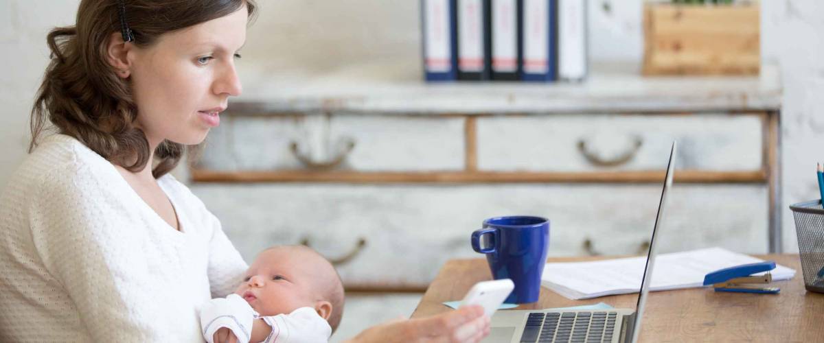 Portrait of young business mom holding her newborn cute babe while looking at cellphone screen.