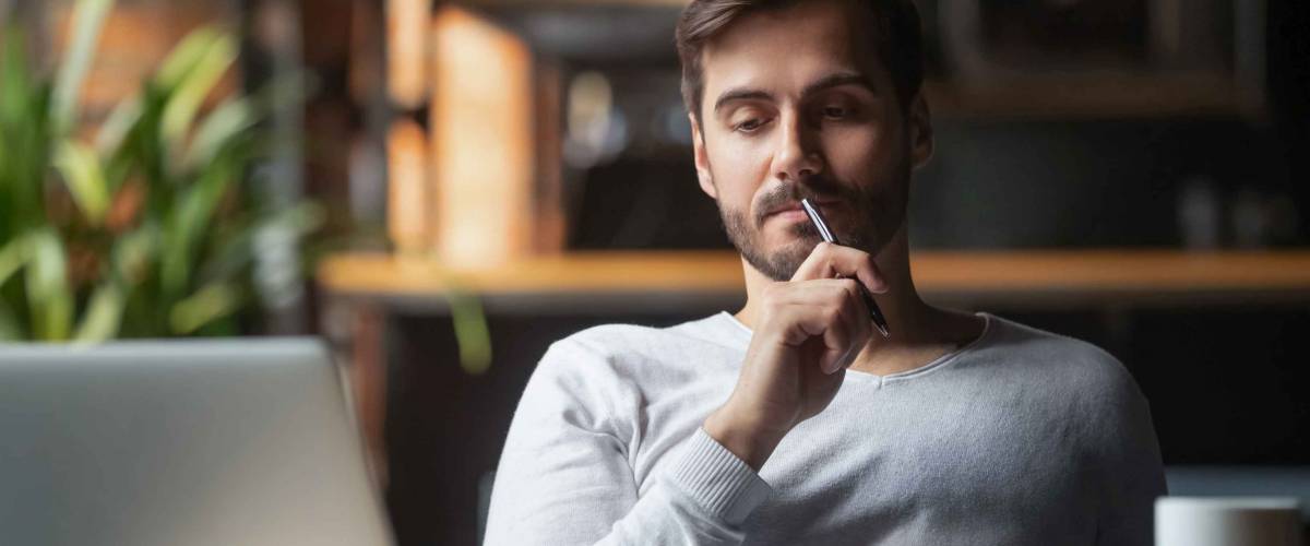 Pensive bearded man sitting at table drink coffee work at laptop thinking of problem solution, thoughtful male employee pondering considering idea looking at computer screen making decision