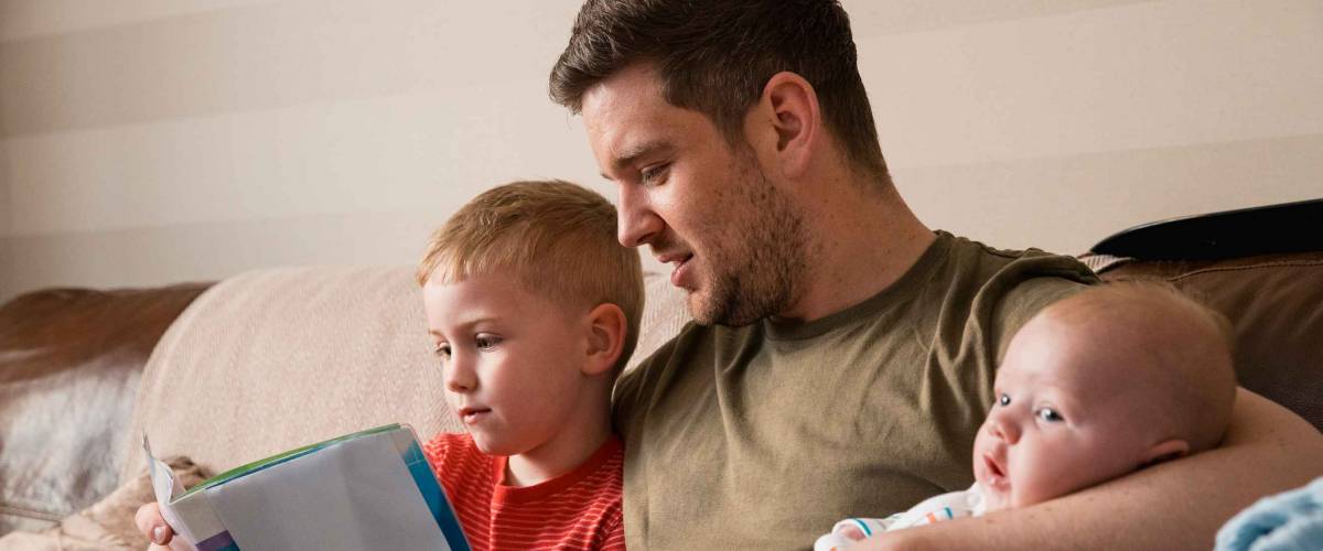 Mid-adult father is holding his newborn baby boy in his arm and then reading though the book with his son under the other arm. They all all sitting on the sofa in the living room.