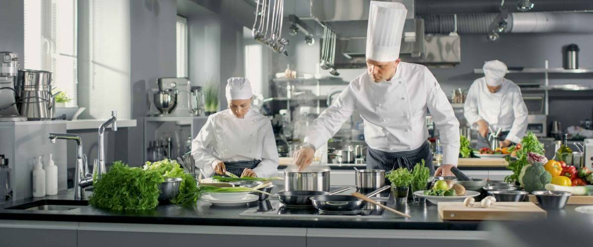 Famous Chef Works in a Big Restaurant Kitchen with His Help. Kitchen is Full of Food, Vegetables and Boiling Dishes.