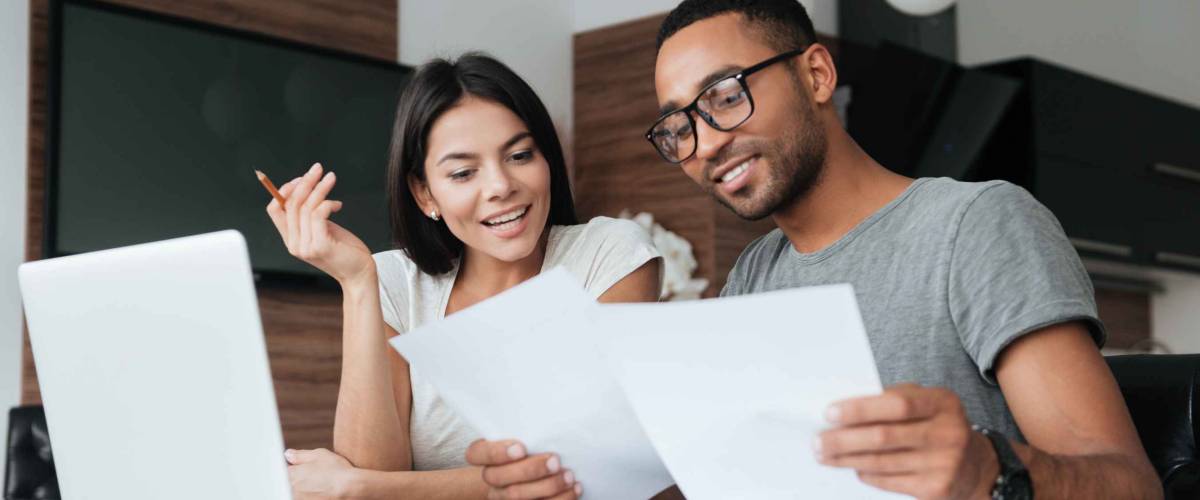 Photo of cheerful loving young couple using laptop and analyzing their finances with documents. Look at papers.