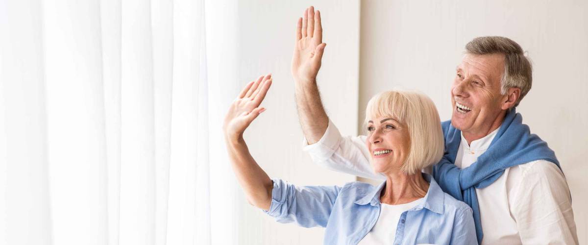 Senior couple waving hands near window, saying goodbye to grandchildren, free space