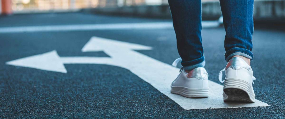 Make decision which way to go. Walking on directional sign on asphalt road. Female legs wearing jeans and white sneakers.