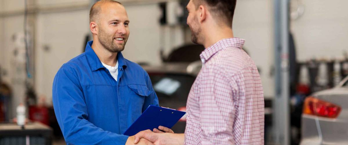 auto service, repair, maintenance, gesture and people concept - mechanic with clipboard and man or owner shaking hands at car shop
