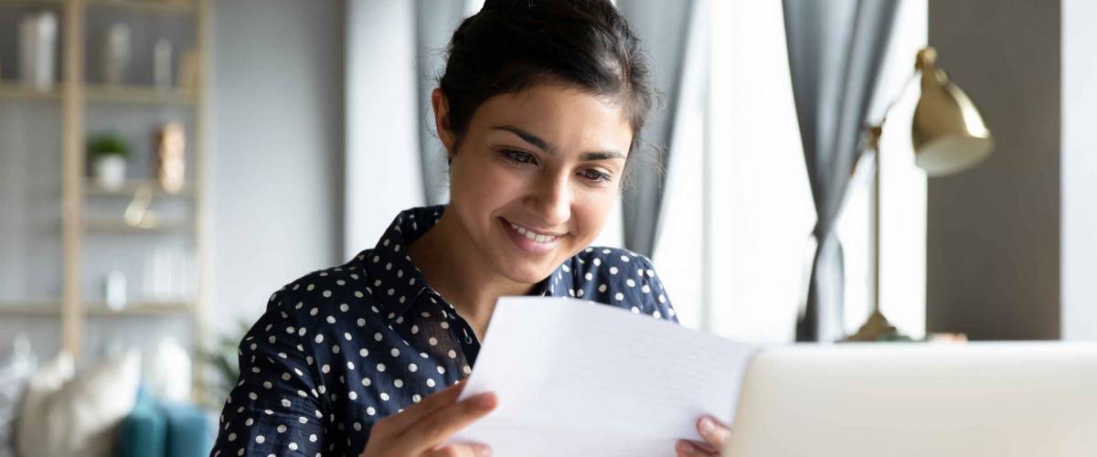 Smiling young woman holding paper bill, sitting in front of laptop.