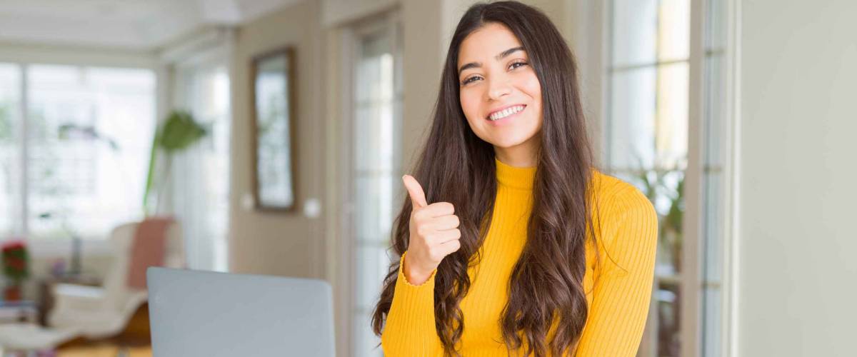 Young woman using computer laptop doing happy thumbs up gesture with hand. Approving expression looking at the camera with showing success.