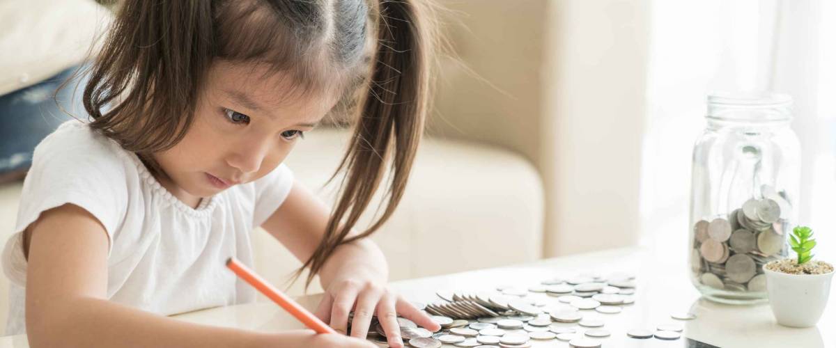 Little girl with pigtails with pile of coins, writing a note on a table.