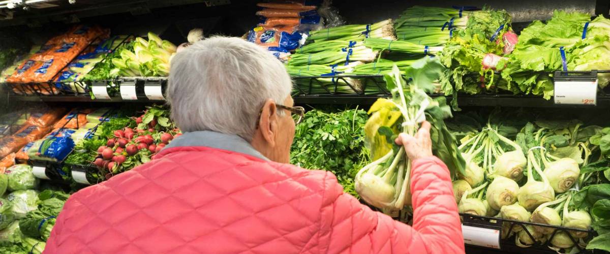 An elderly, gray-haired woman in a pink coat shopping for kohlrabi in the produce section of a market.
