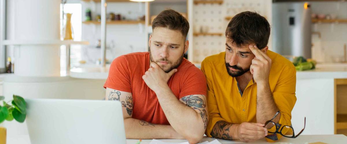 A gay couple paying bills online. They are talking about how much they spent on unnecessary things.