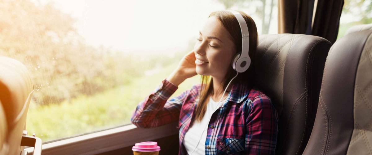 The woman on the passenger seat of the bus listens to music and drinks coffee. She looks out the window and smiles. Outside the window is a beautiful green landscape.