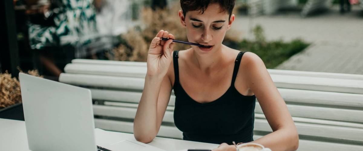 Young woman chews pen while looking down at cellphone in her hand.