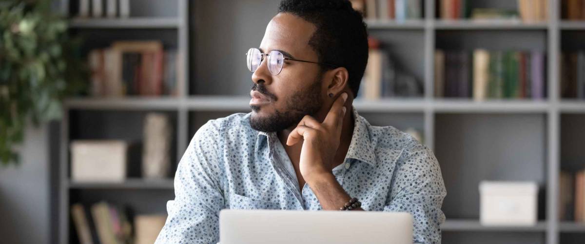 Dreamy thoughtful African American businessman looking to aside, sitting at work desk with laptop, serious man pondering project plan or strategy, making difficult decision, solving problem