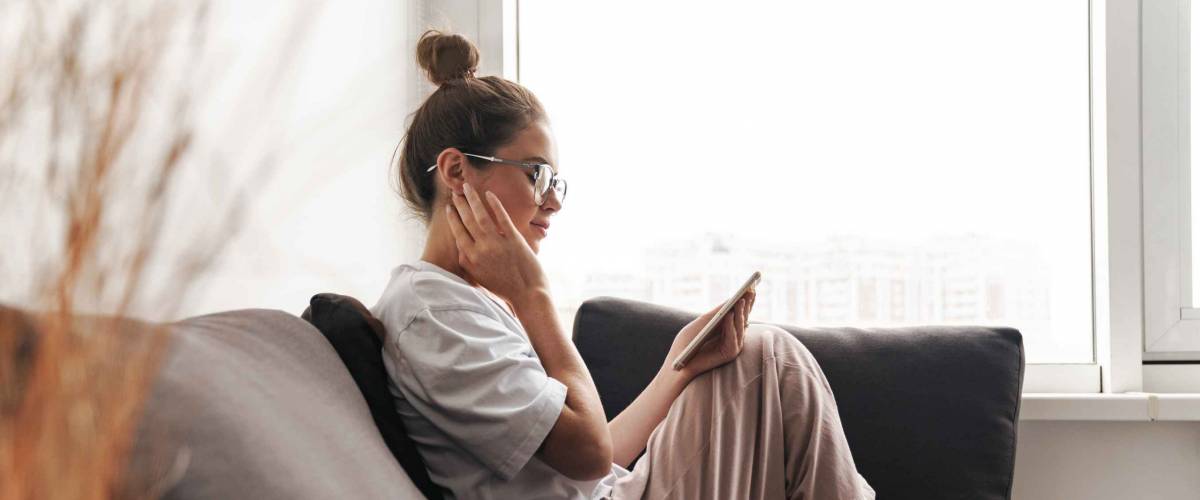 Image of concentrated nice woman in eyeglasses using cellphone while sitting on sofa at living room