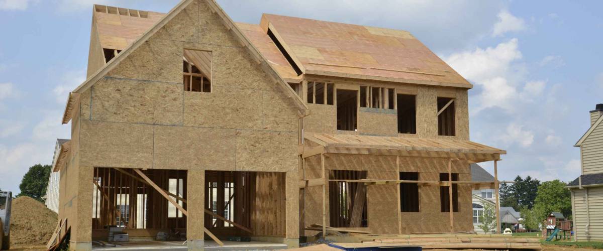 A single family home under construction. The house has been framed and covered in plywood. There is a two car garage with a gravel driveway.