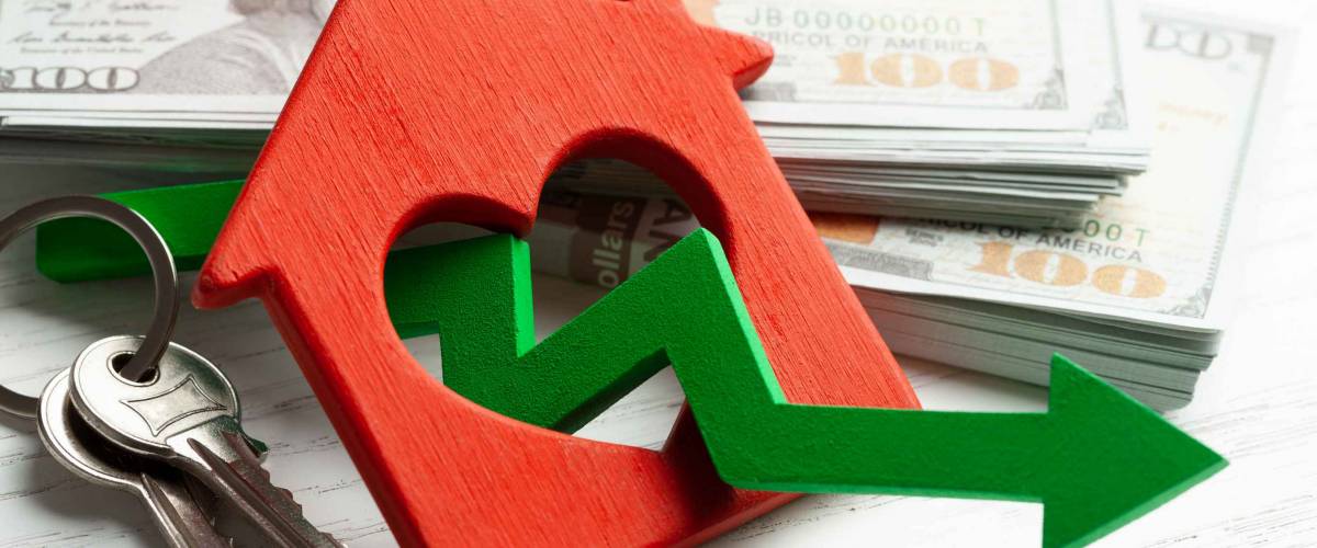 Arrow down and house with money on white wooden background. Falling market price of real estate.