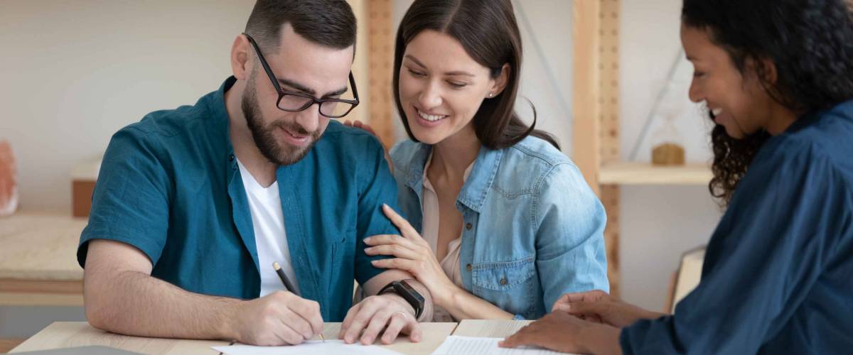 Smiling young family clients make purchase deal sign insurance contract at meeting with african american estate agent. Happy millennial couple reading document about buying house on loan.