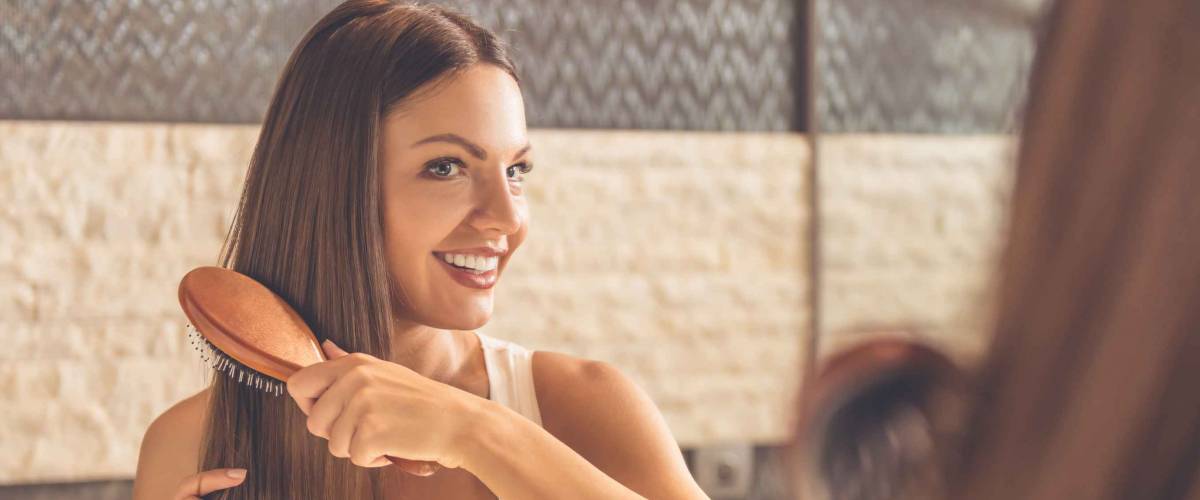Beautiful young woman in white undershirt is combing her hair and smiling while looking into the mirror in bathroom