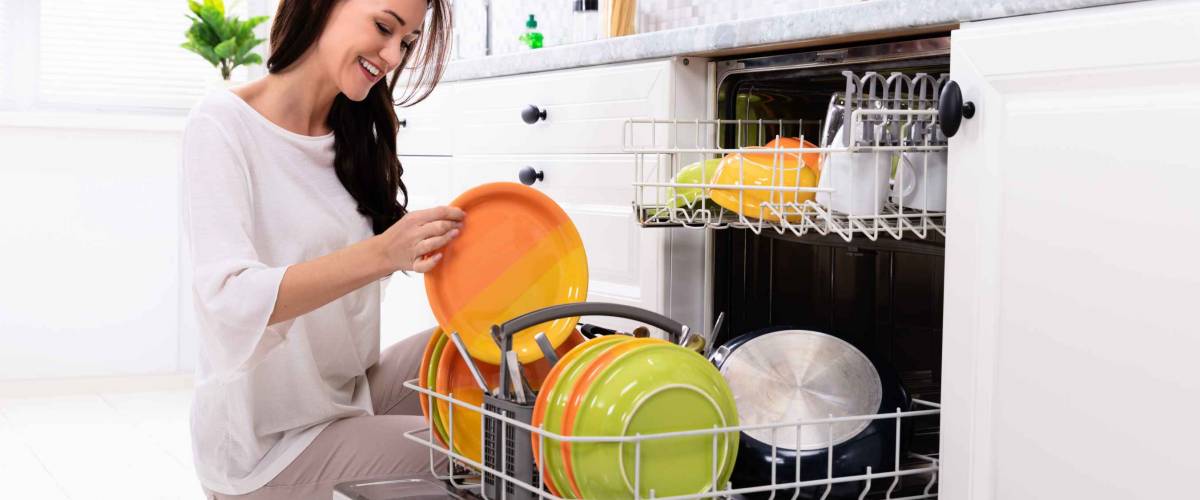 Smiling Young Woman Arranging Plates In Dishwasher At Home