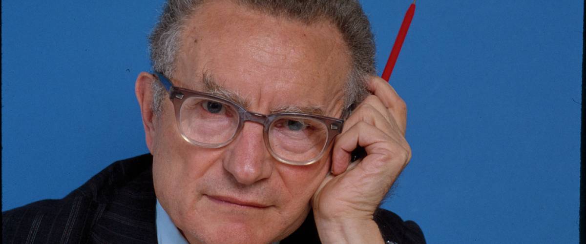 Paul A. Samuelson looks at camera with pen in his hand, posed at the side of his head.