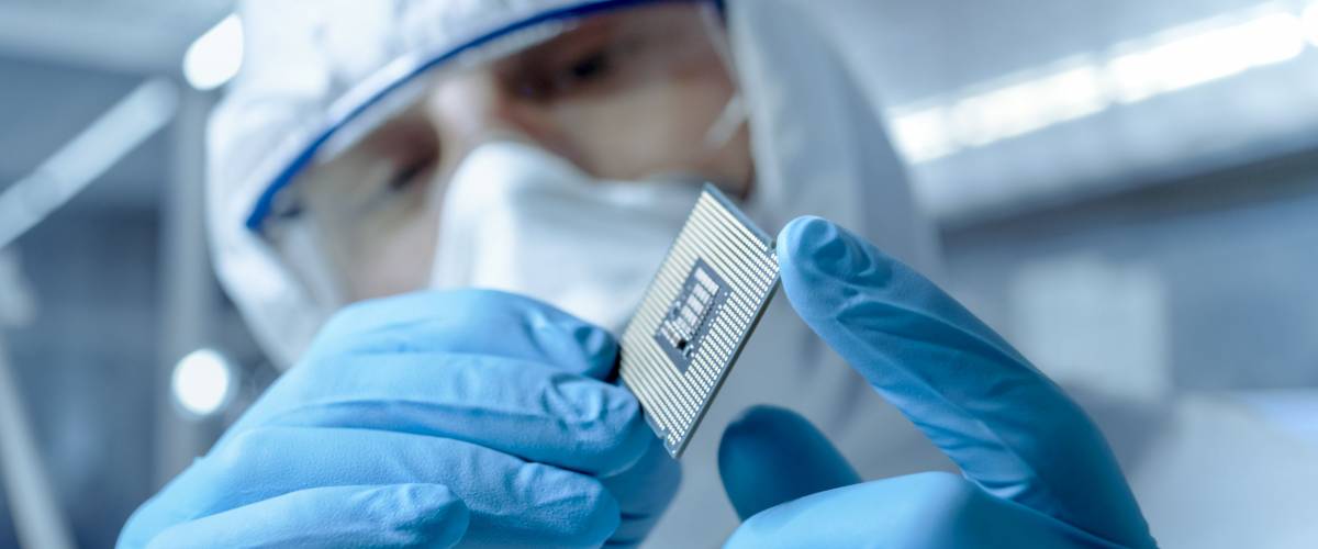 In Ultra Modern Electronic Manufacturing Factory Design Engineer in Sterile Coverall Holds Microchip with Gloves and Examines it.
