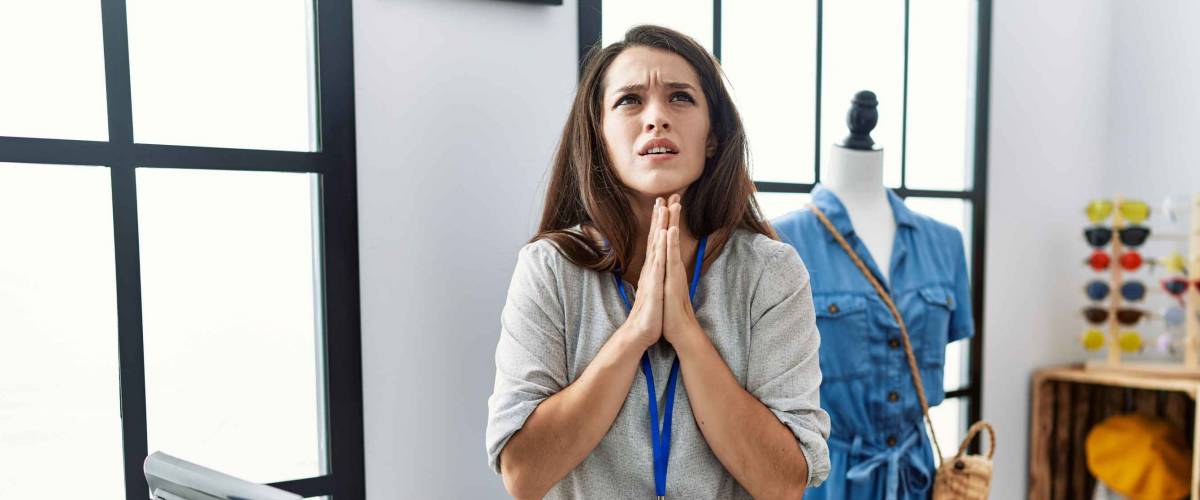 Young brunette woman working as manager at retail boutique begging and praying with hands together with hope expression on face very emotional and worried. begging.