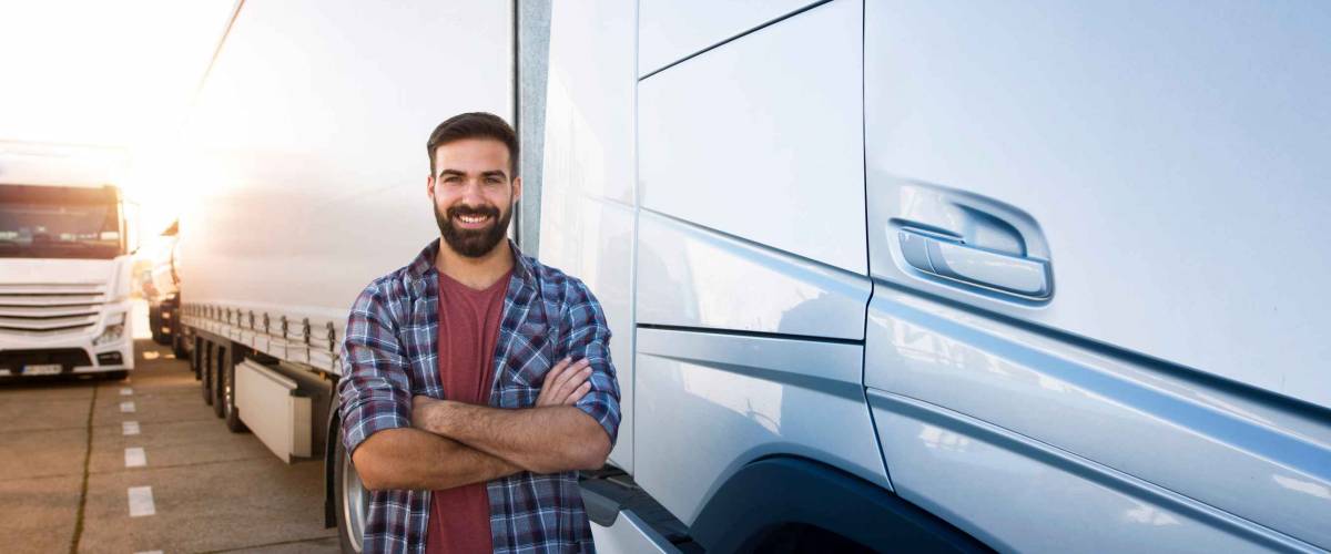 Portrait of young bearded man standing by his truck. Professional truck driver with crossed arms standing by semi truck vehicle.