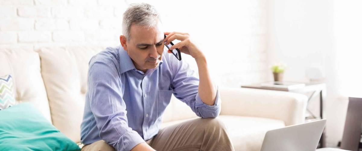 Busy mature man sitting in living room talking on mobile phone and making notes.