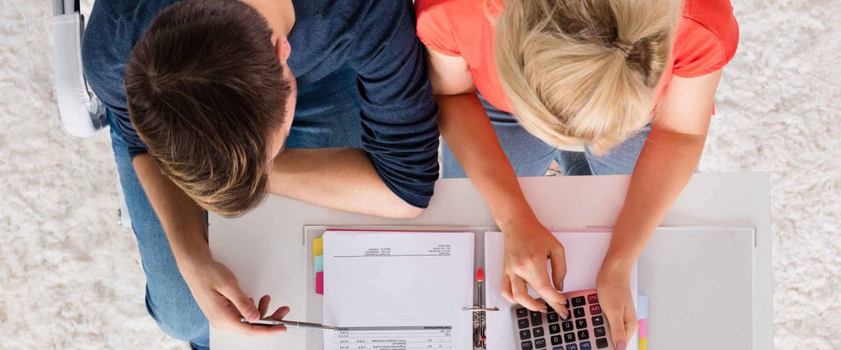 Couple Using Calculator For Calculating Bills On Desk At Home. Seen From Above.