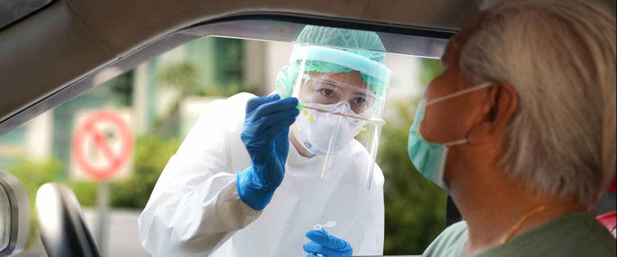 A nurse in a face shield and mask uses a long swab to test a man in his car for COVID-19
