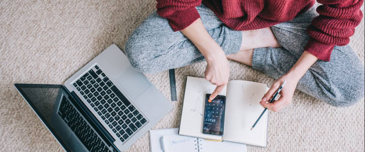 A person in sweatpants sitting cross-legged on the carpet reviewing their budget with a phone calculator