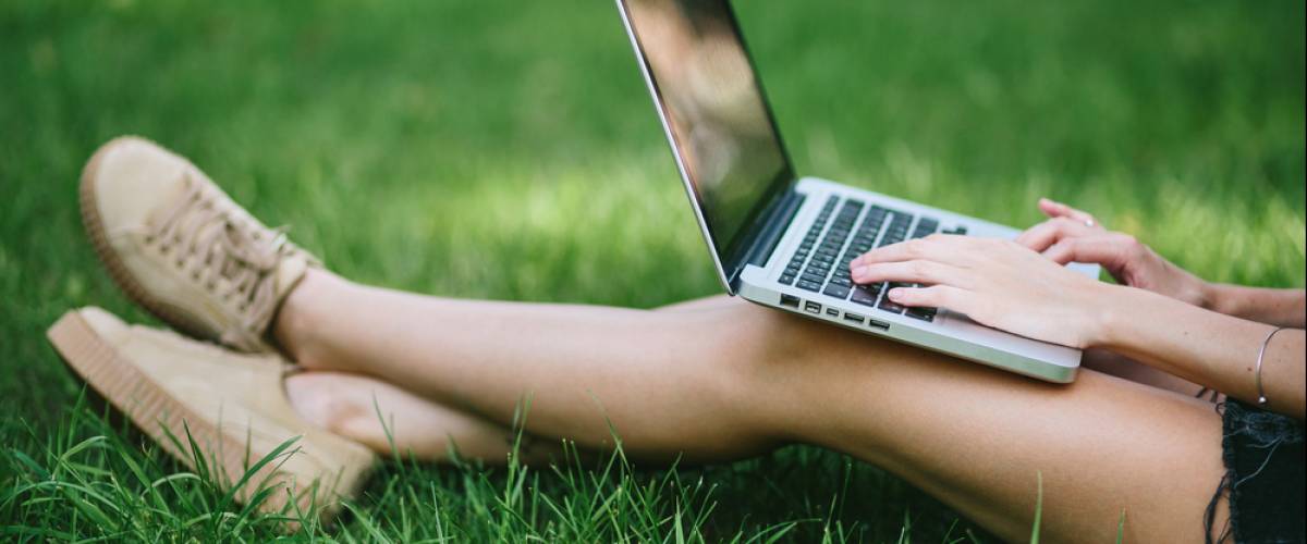 A person wearing shorts and tennis shoes sitting in the grass with their ankles crossed and using a laptop