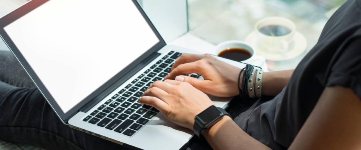 A person types on the computer while sitting on a windowsill with a cup of coffee