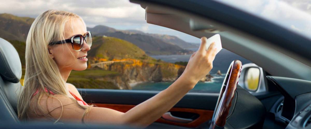 travel, road trip and people concept - happy young woman in convertible car taking selfie by smartphone over big sur coast of california background