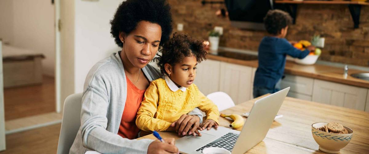 Black working mother taking notes while daughter is sitting on her lap and using laptop at home. Small boy is in the background.