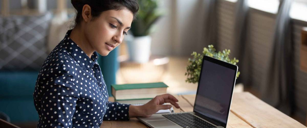 Millennial Indian girl sit at desk in living room study on laptop making notes.