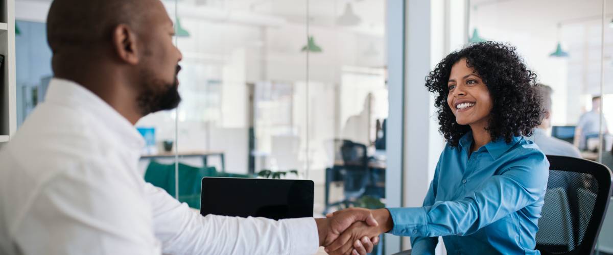 Smiling African American manager sitting at his desk in an office shaking hands with a job applicant after an interview