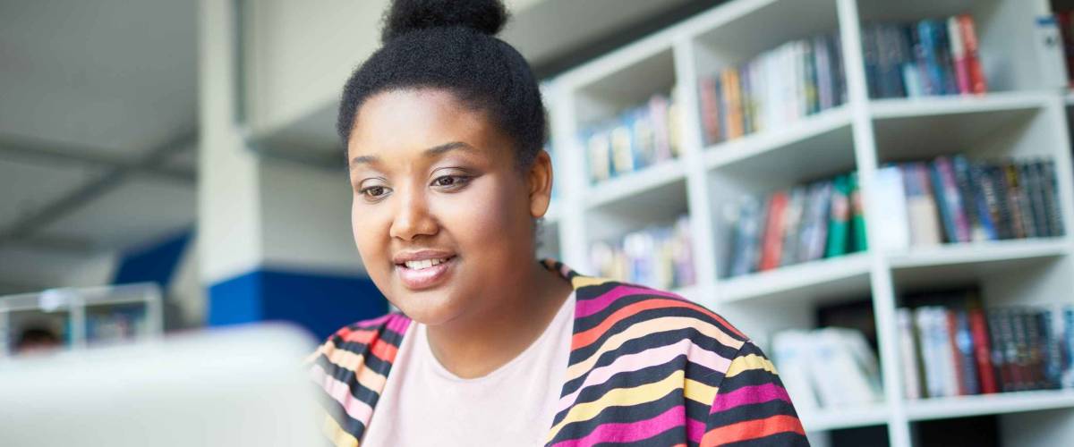Cheerful confident young black, curvy student reading online article on laptop while preparing for exam in library
