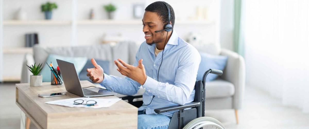 Disabled black guy with headset communicating online on laptop, having business meeting from home. Millennial African American man in wheelchair working in call centre remotely