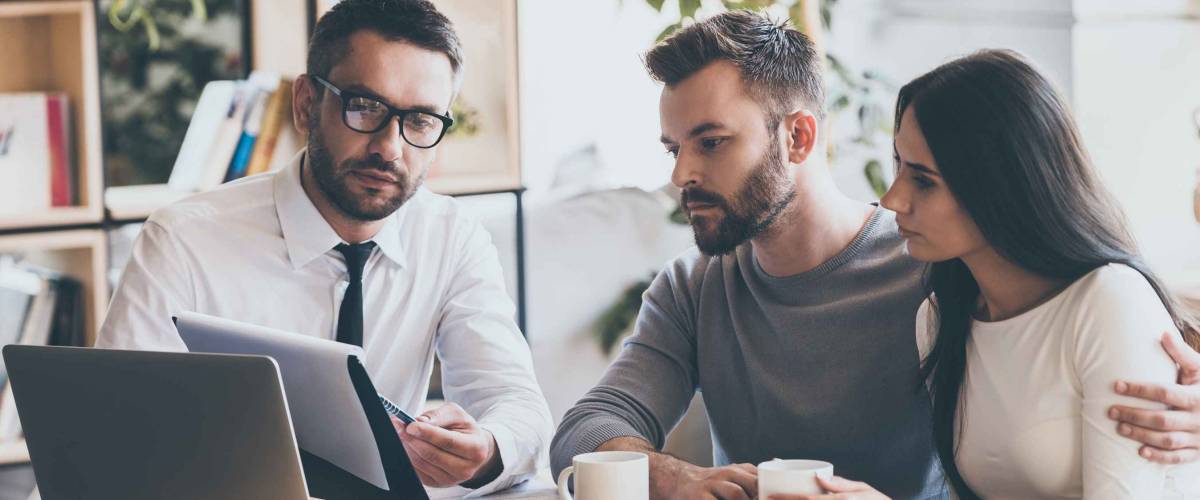 I need your signature here. Confident young man in shirt and tie holding some document and pointing it while sitting together with young couple at the desk in office