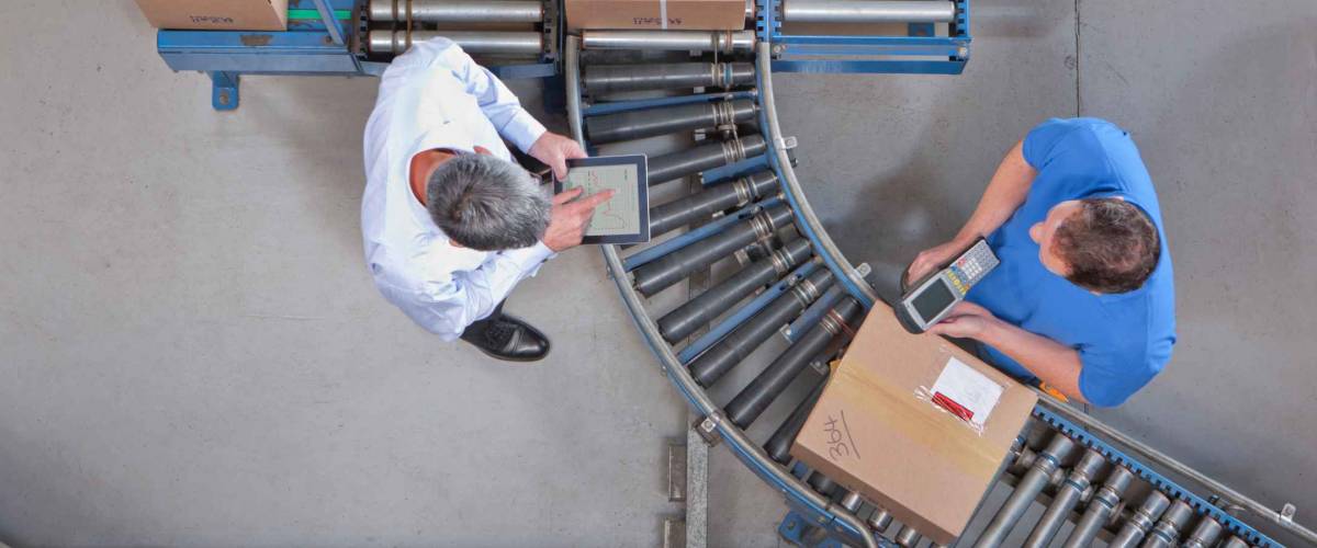 overhead view of two white men, one worker and the other a manager, in a factory surrounded by wheeled conveyor belt
