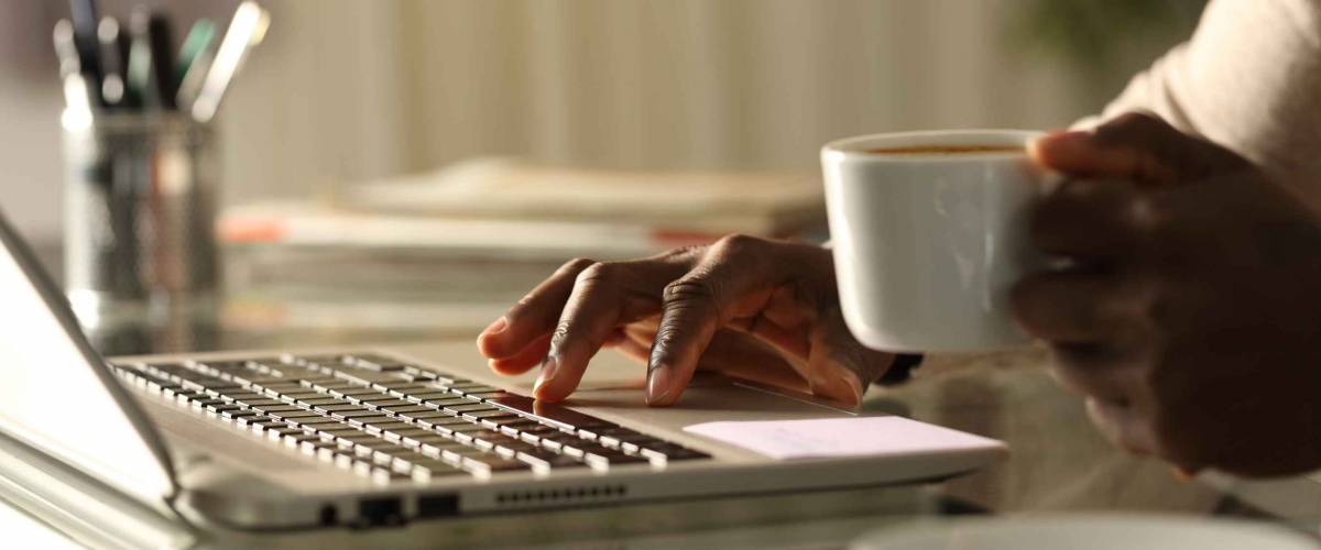 Close up of a black man hands using laptop holding a coffee cup in the night on a desk at home