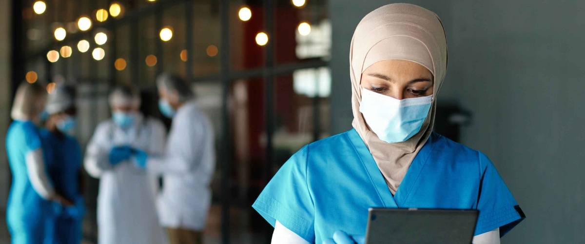 Young hijabi woman doctor in medical mask and gloves standing in hospital and using tablet device.