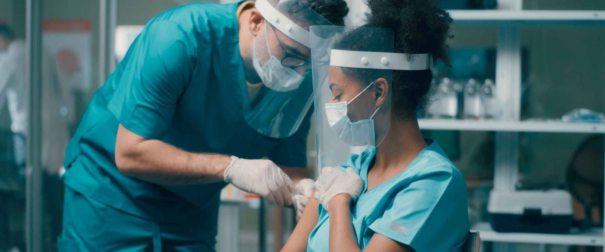 Male medical practitioner making vaccine injection to ethnic female colleague while working in hospital together