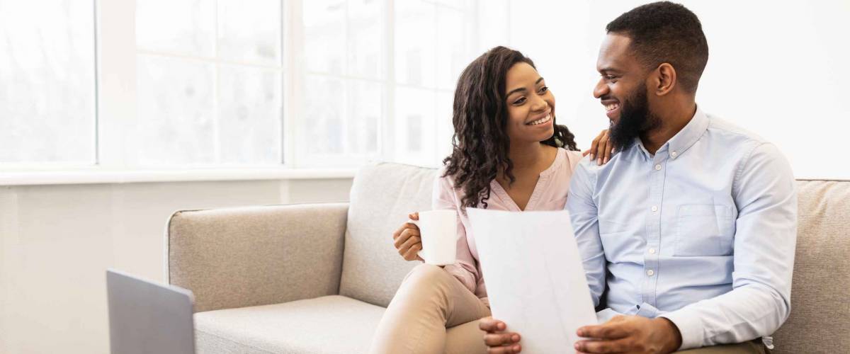 Planning Family Budget. Smiling african american man holding paper, working and reading documentation at home, woman drinking coffee and looking at him, sitting on the couch with laptop