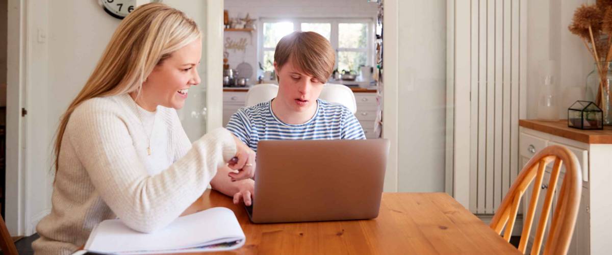 Downs Syndrome Man Sitting With Home Tutor Using Laptop For Lesson At Home