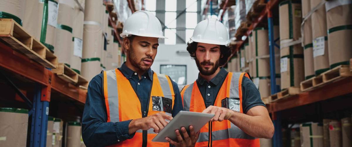 Focused manager and employee standing in warehouse discussing inventory stock while standing between cardboard packed goods with safety hardhat and vest